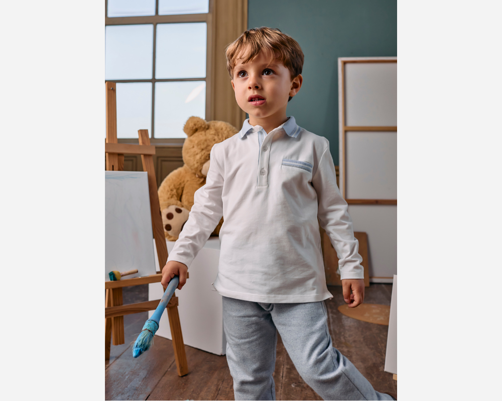 Child in a light-colored shirt and pants holding a blue toy broom in a room with a teddy bear and chair.